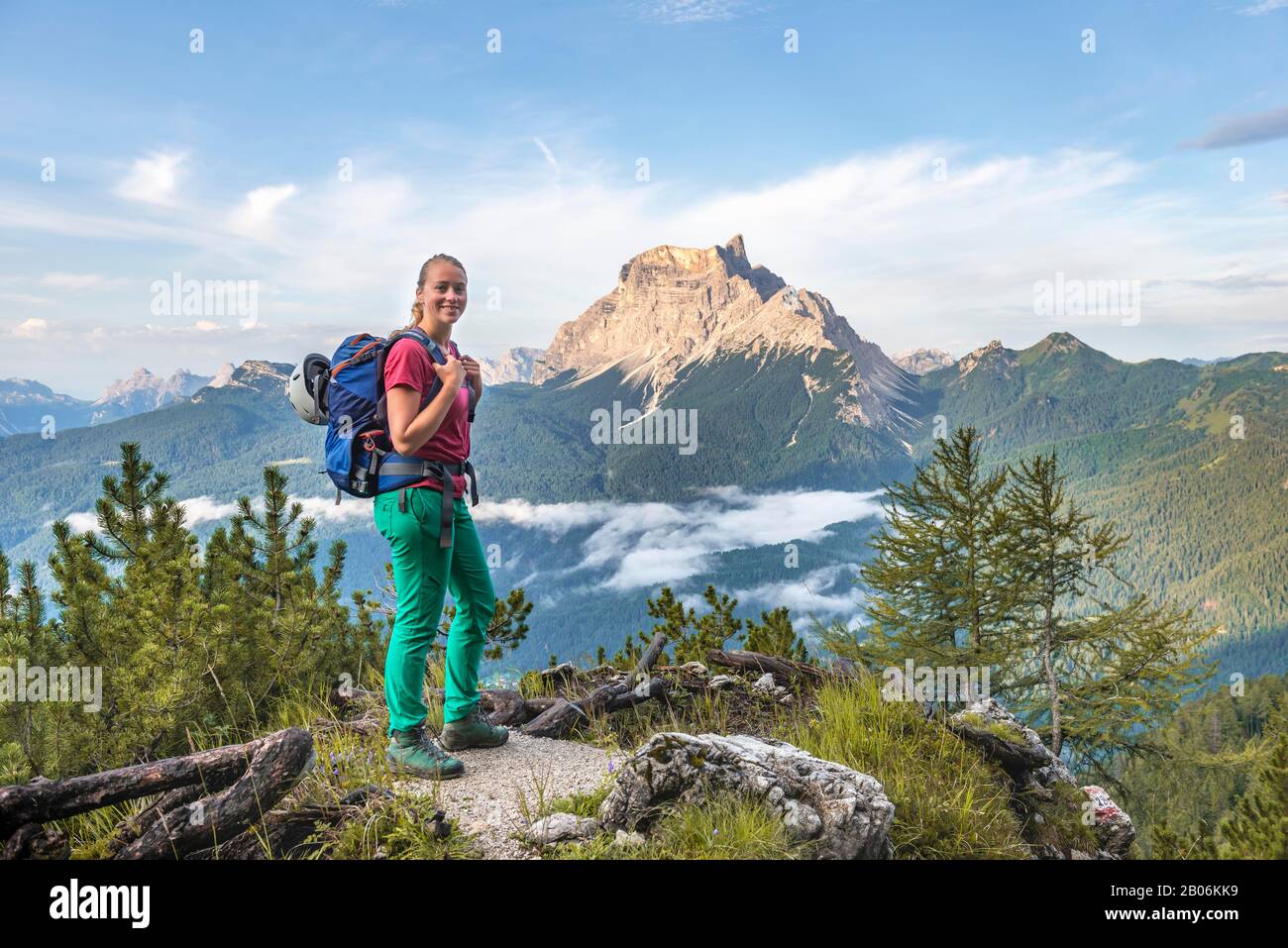 Giovane escursionista femminile guarda alla macchina fotografica, dietro la sua vetta di montagna la Rocheta, Dolomiti, Belluno, Italia Foto Stock