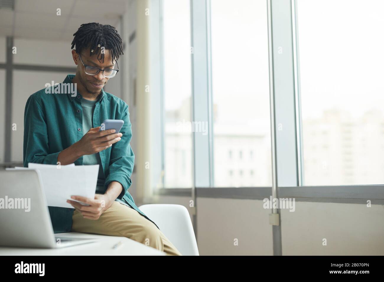 Giovane uomo d'affari africano in occhiali seduti sul tavolo leggendo un contratto e utilizzando il suo cellulare nel lavoro Foto Stock