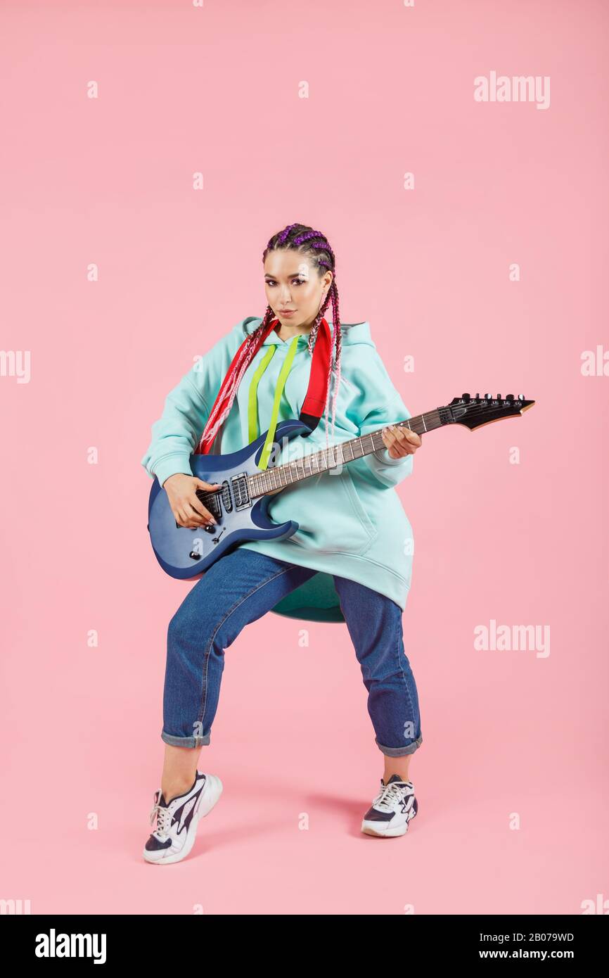Giovane chitarrista donna in abiti oversize con chitarra elettrica isolata su sfondo rosa. Foto Stock