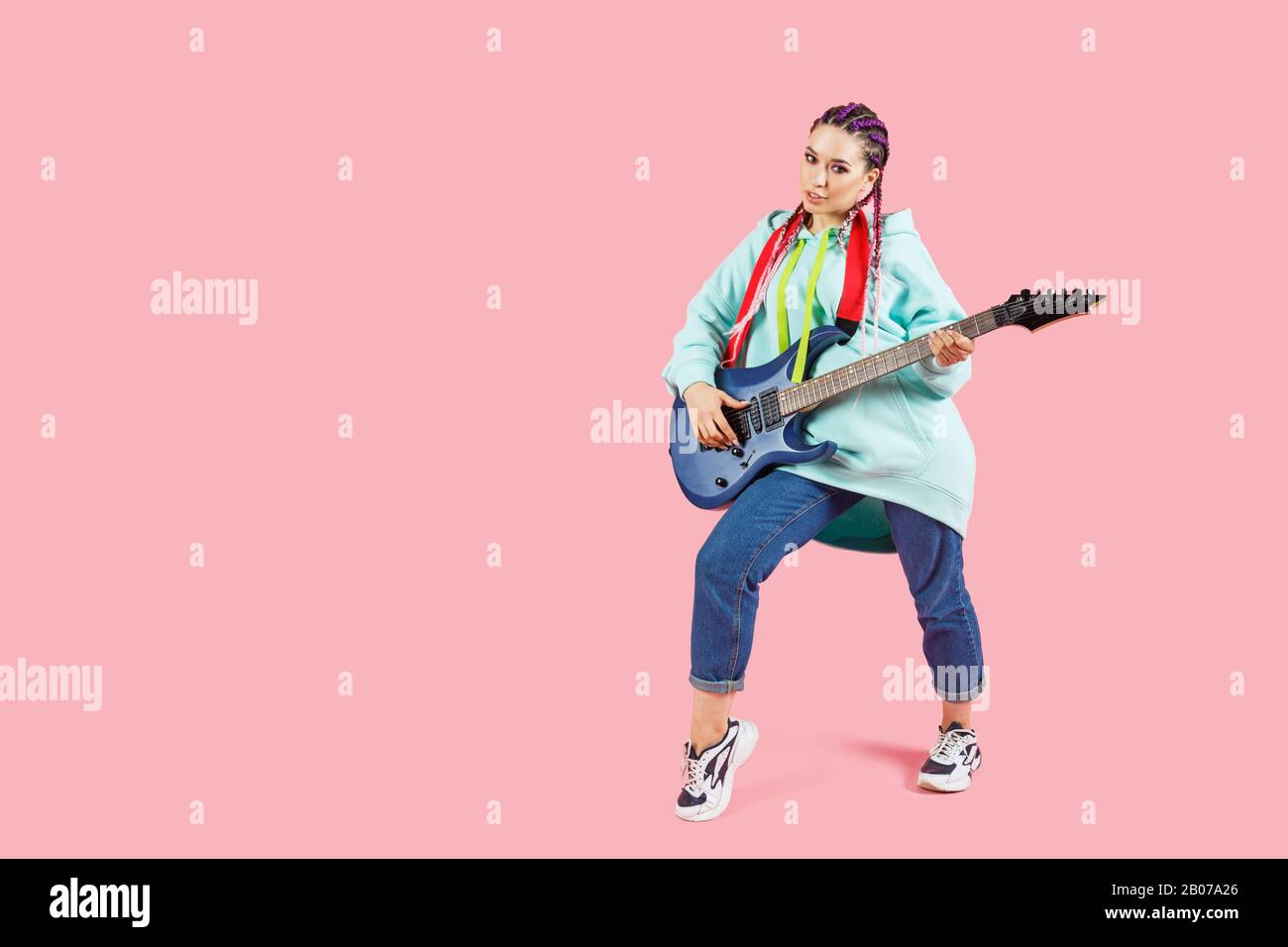 Giovane chitarrista donna in abiti oversize con chitarra elettrica con copyspace isolato su sfondo rosa Foto Stock