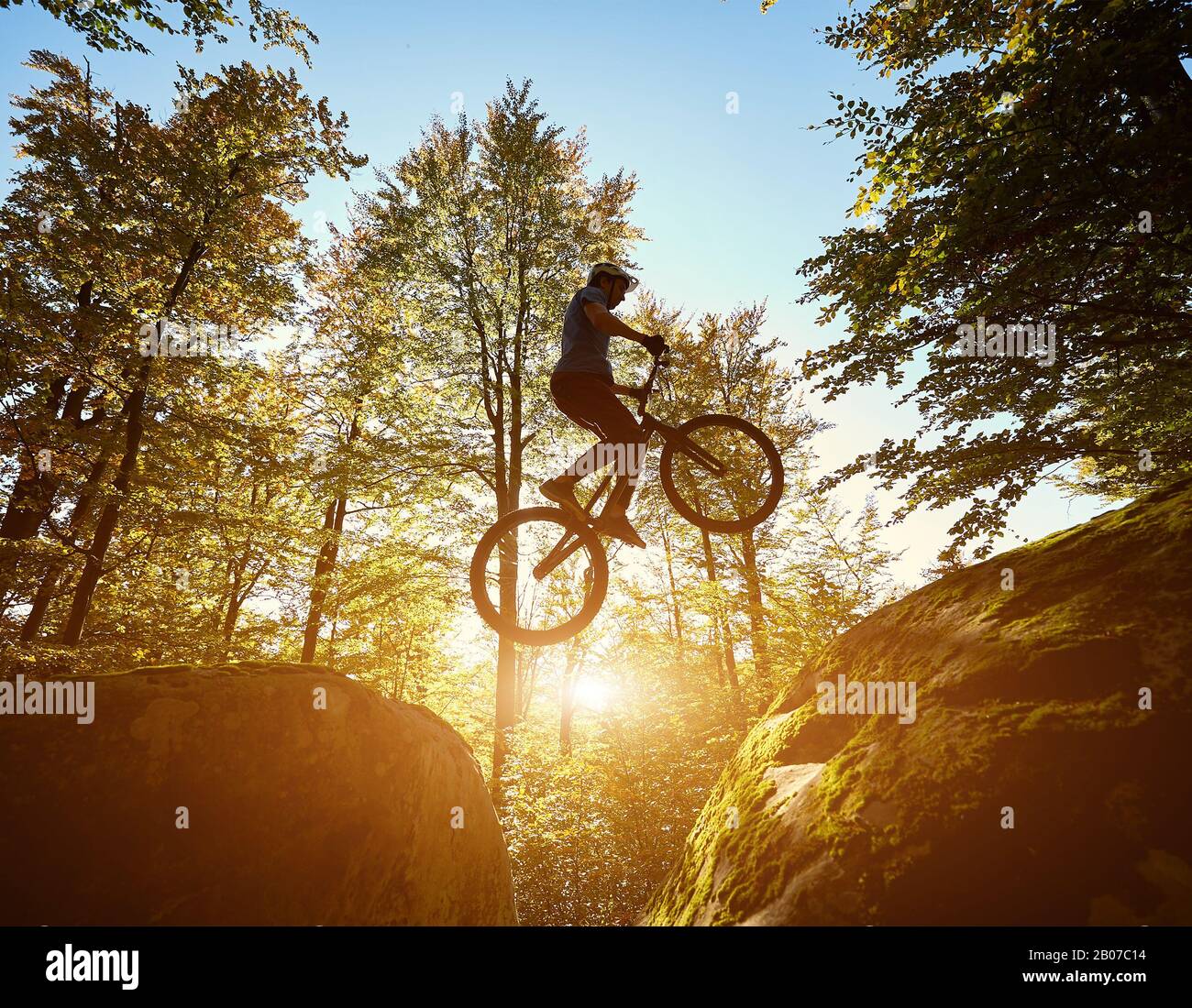 Giovane ciclista maschile che salta in bicicletta di prova tra due grandi massi, pilota professionista che fa acrobatico trucco nella foresta in giornata di sole. Concetto di sport estremo stile di vita attivo Foto Stock