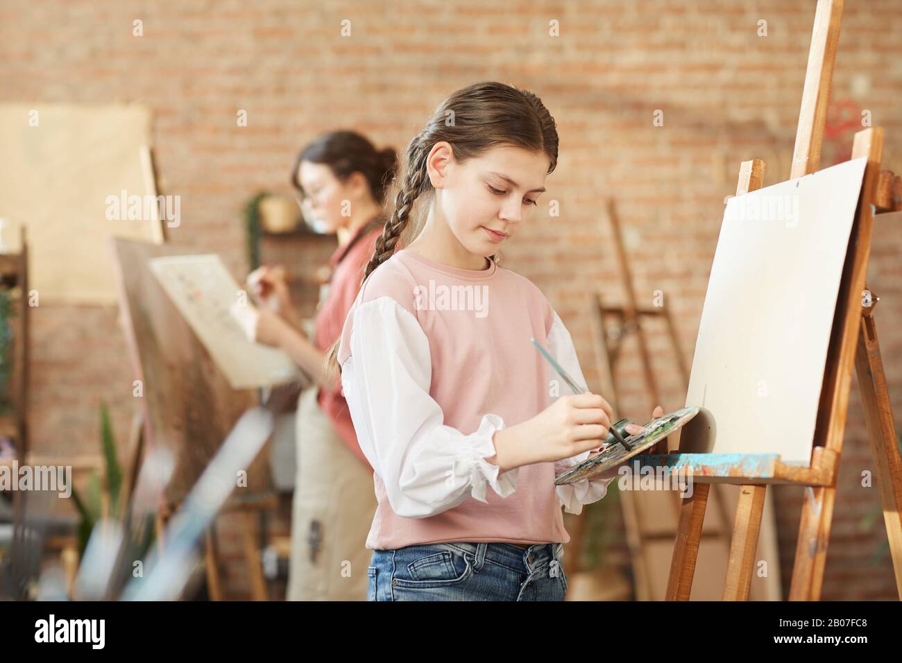 Giovane ragazza in abbigliamento casual imparare a dipingere su cavalletto durante la sua lezione in studio d'arte Foto Stock
