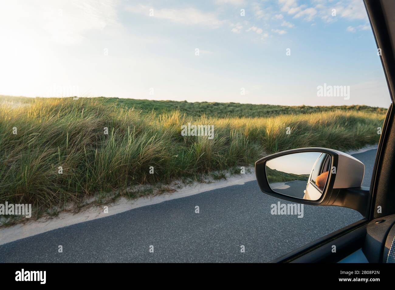 Guida sul concetto dell'isola di Sylt, in Germania. Finestra auto vista del paesaggio di dune di erba marram e una strada sabbiosa. Specchietto retrovisore per auto. Foto Stock