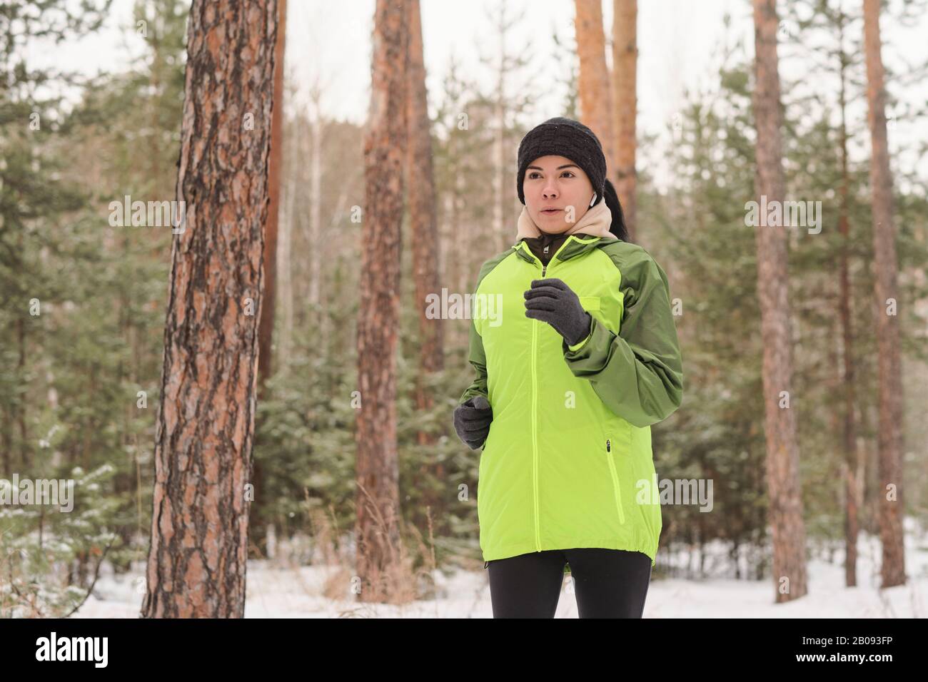 Ragazza sportiva con giacca verde che corre da sola nel parco forestale invernale mentre si allenano all'aperto Foto Stock