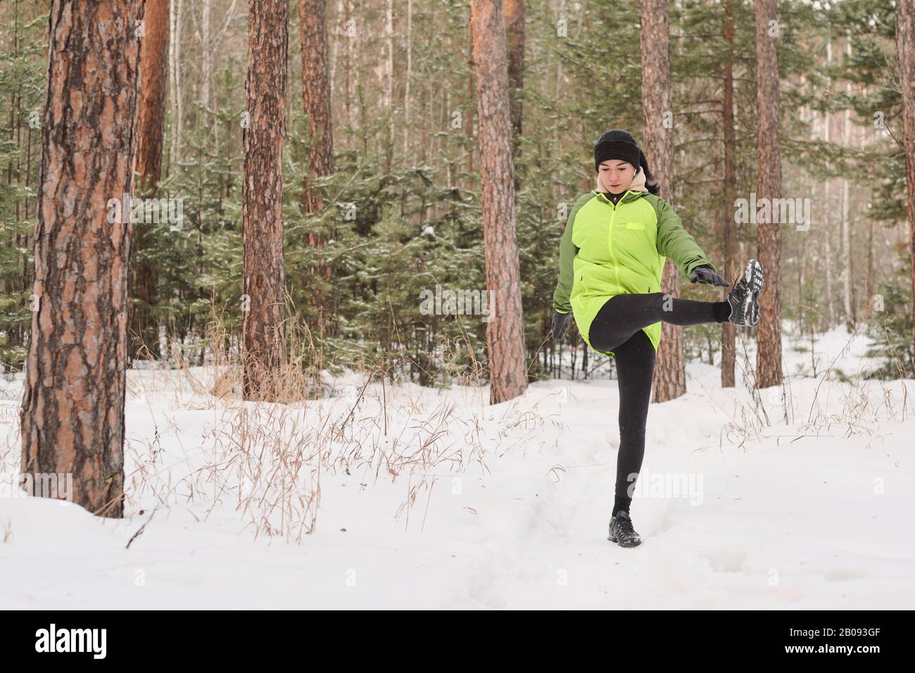 Giovane donna in giacca verde in piedi nella foresta invernale e sollevamento gamba da parte durante l'allenamento Foto Stock