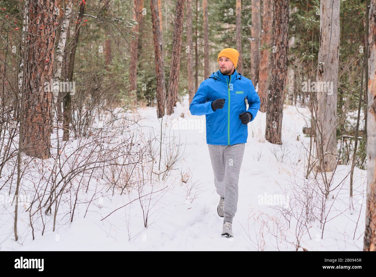 Bel giovane uomo in cappello che corre su strada innevata in bella foresta mentre allenarsi da solo Foto Stock