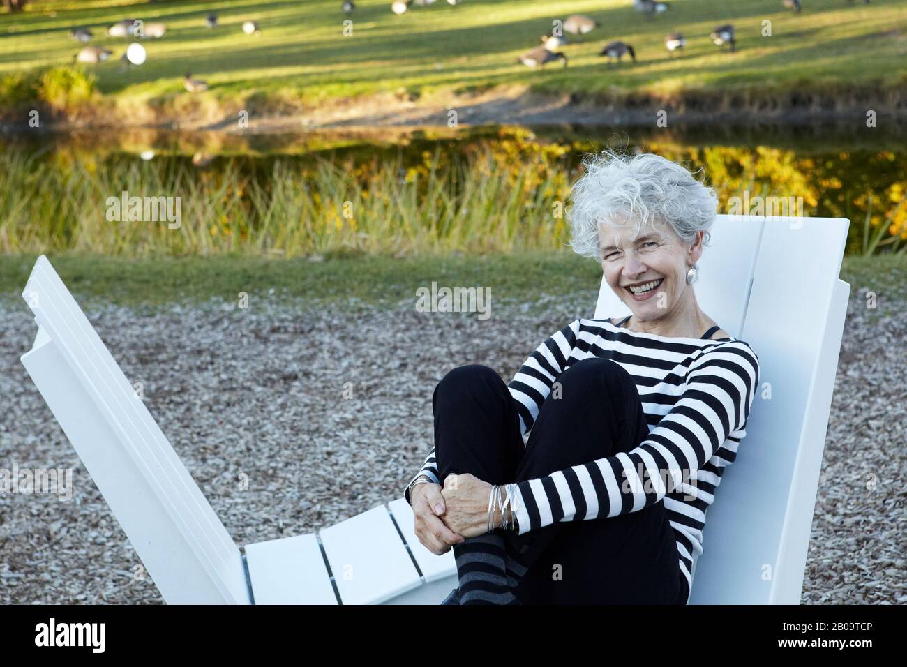Bella, felice Senior donna seduta sulla sedia Adirondack nel parco con stagno e oche Foto Stock