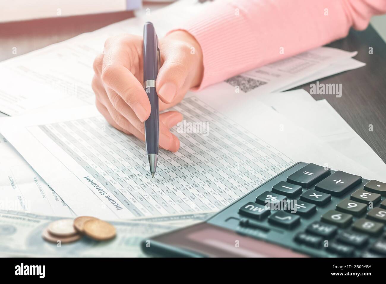 Closeup vista laterale sulla mano di contabile femminile che sta tenendo una penna e lavorando in un ufficio con una pila di documenti e tabelle. Calcolo di Foto Stock
