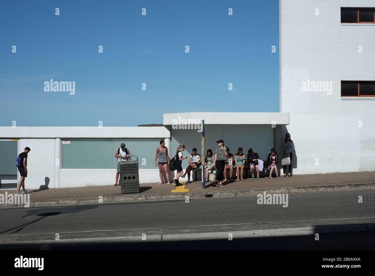 Persone in attesa di una fermata del bus Bondi Beach; edificio bianco tagliato contro un cielo blu in una giornata di sole, su Campbell Parade, Sydney, Australia Foto Stock