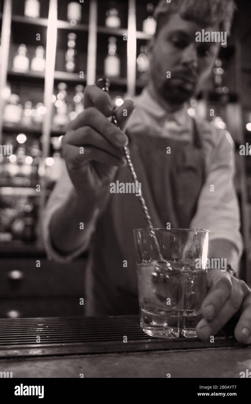 Barman che mescola un vecchio stile sul bar con un bastoncino molto lungo, in vetro, testa in colpo a destra - camicia bianca, grembiule, bottiglie sugli scaffali Foto Stock
