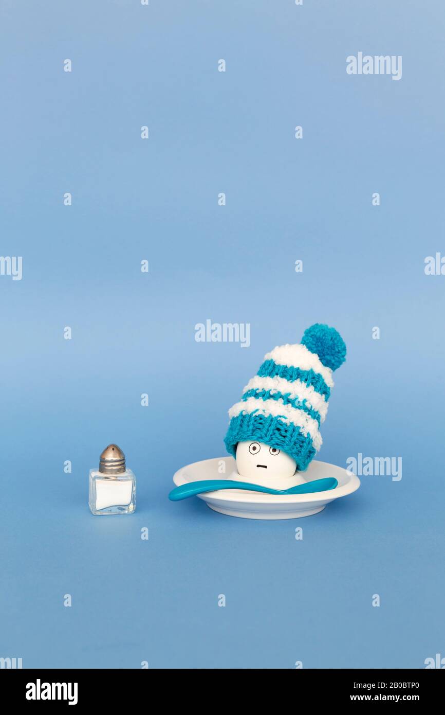 Divertente concetto di colazione con un uovo bollito con astratto grumpy volto umano indossando un cappello di lana lavorato a maglia su sfondo blu Foto Stock