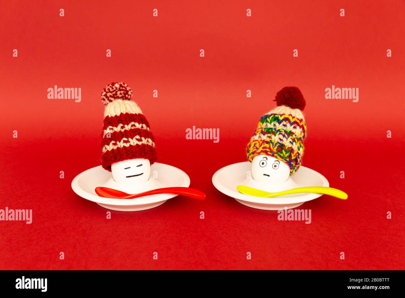 Divertente concetto di colazione con uova sode con astratto felice e infastidito volto umano indossando cappelli di lana lavorato a maglia su sfondo rosso Foto Stock
