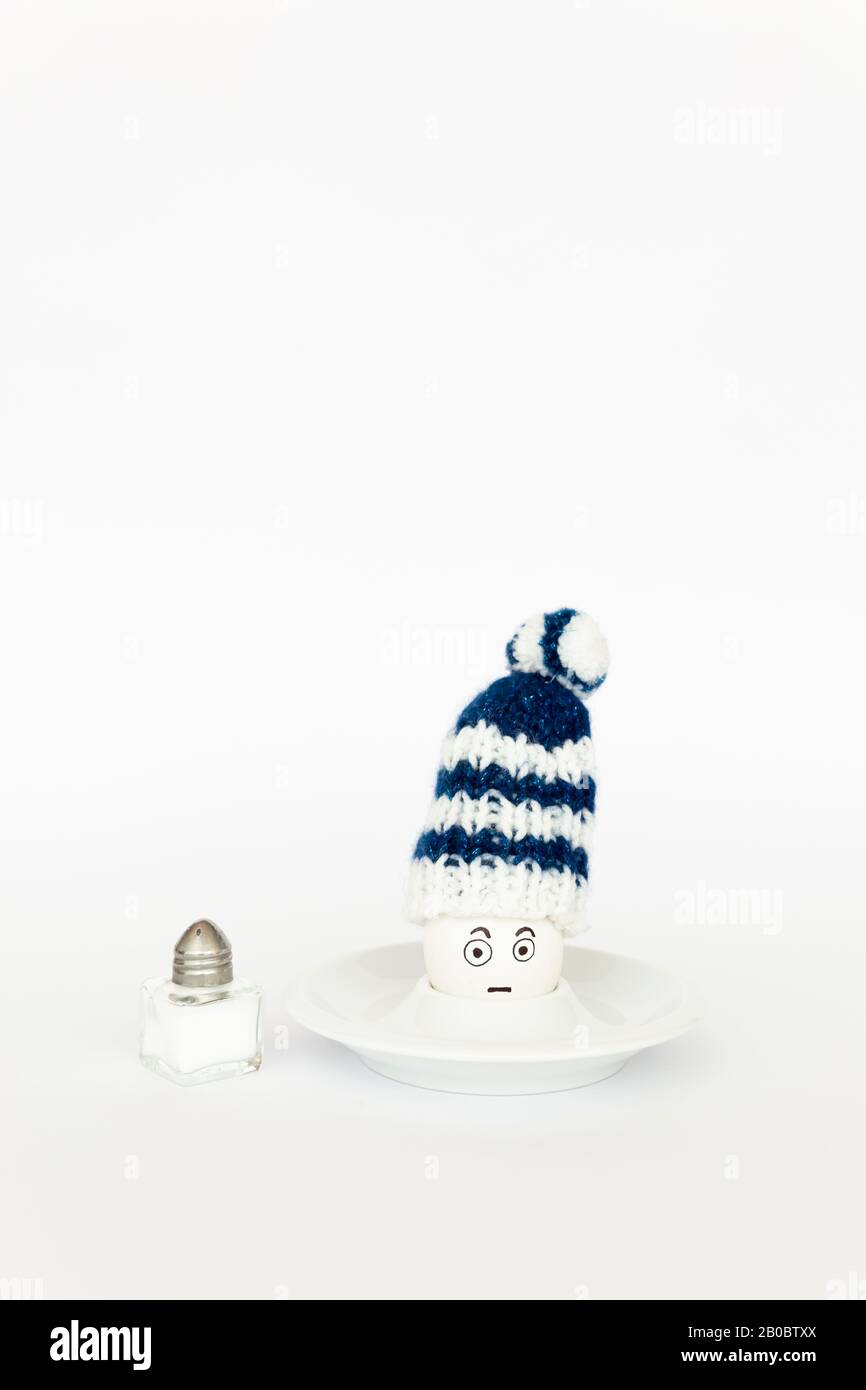 Divertente concetto di colazione con un uovo bollito con astratto grumpy volto umano indossando un cappello di lana lavorato a maglia su sfondo bianco Foto Stock