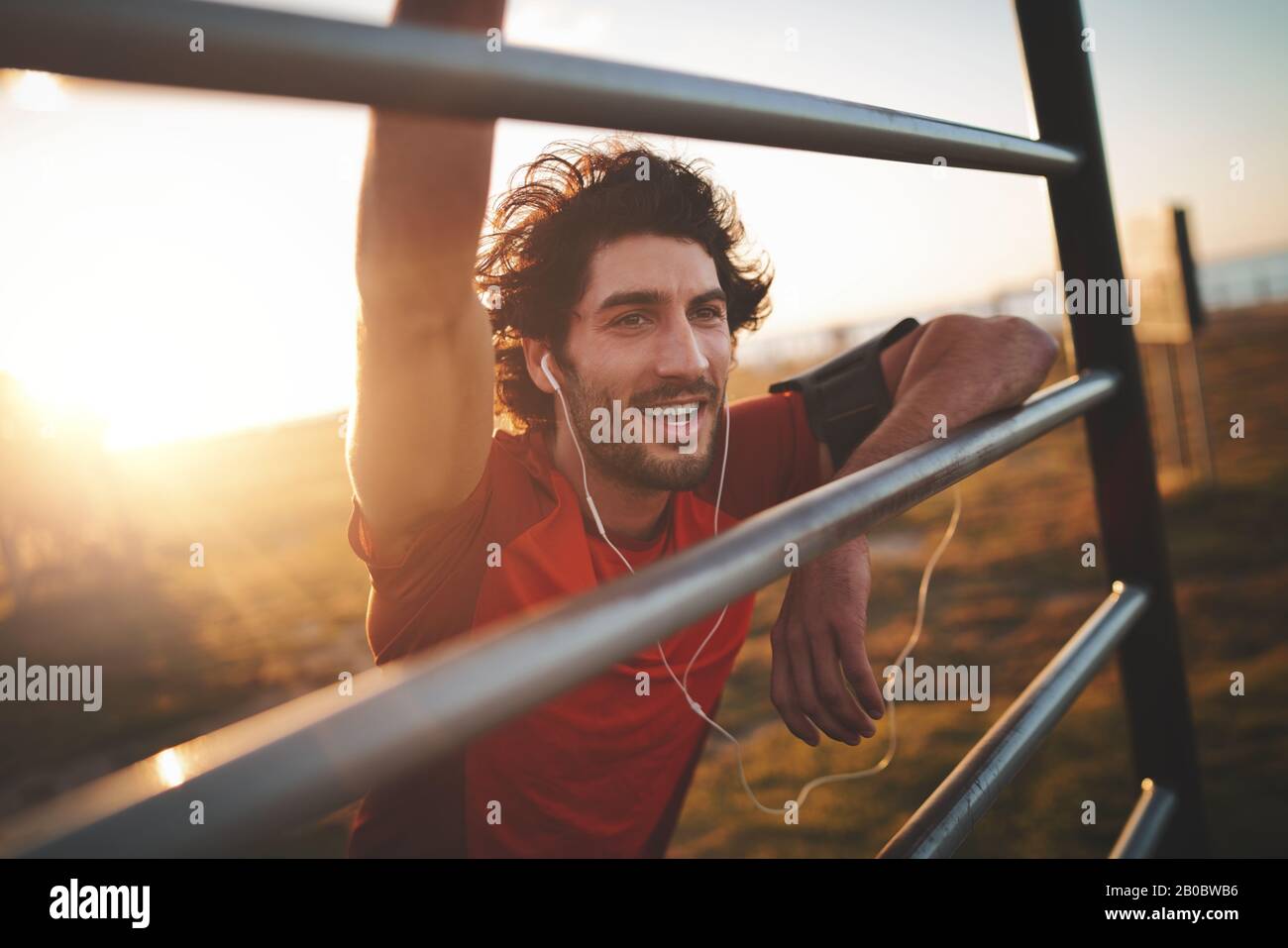 Ritratto di un giovane e allegro corridore maschio con gli auricolari nelle orecchie appoggiati sulla ringhiera dopo l'esercizio nel parco in una giornata di sole Foto Stock
