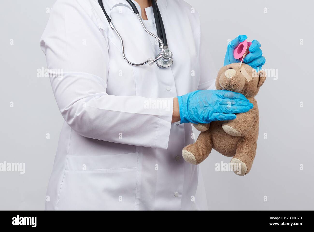 medico femminile in un camice bianco e guanti di lattice blu tiene un orsacchiotto marrone senza occhio, medico avvolge la testa con cerotto adesivo Foto Stock