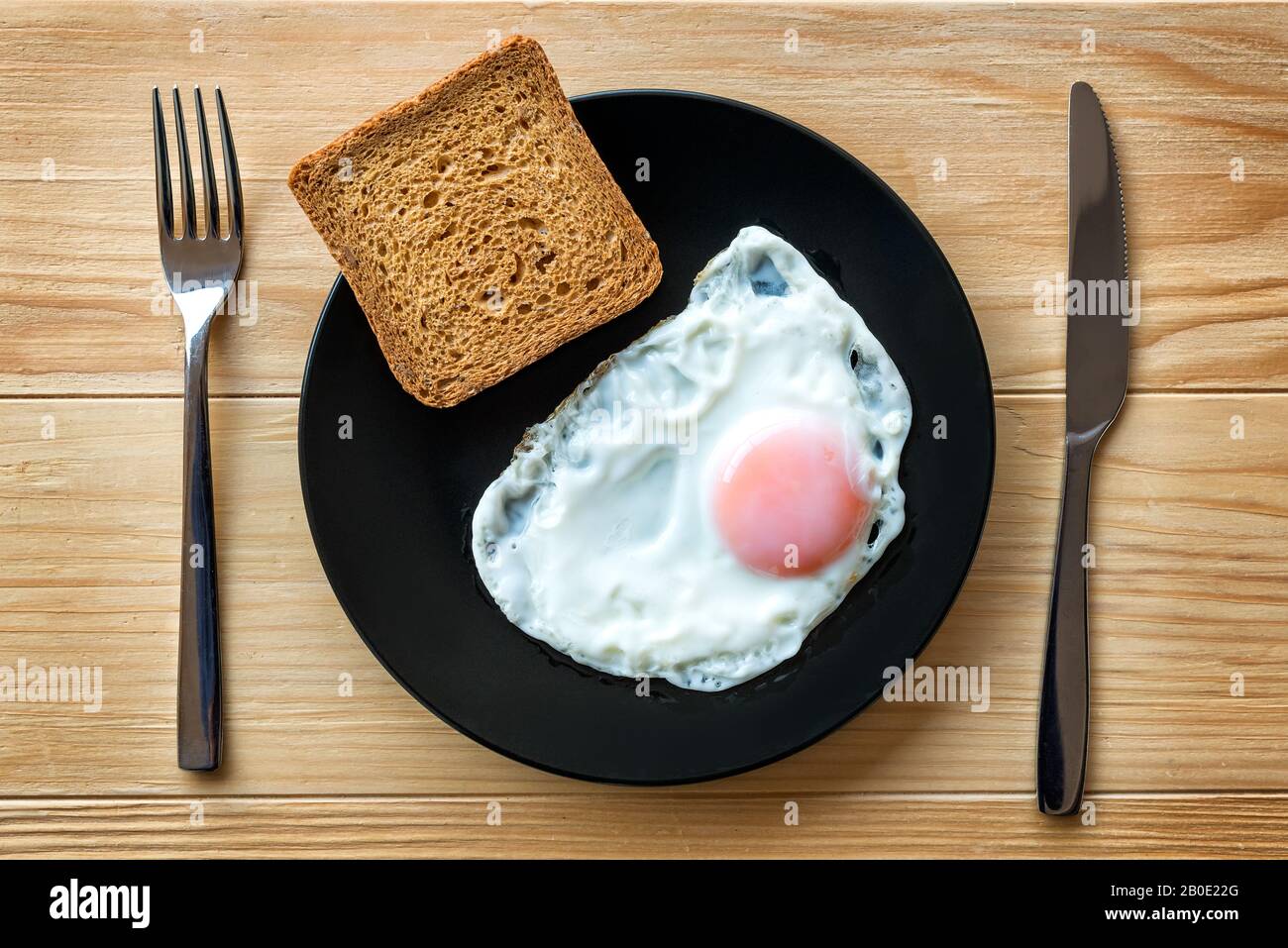 Uovo fritto chiudere in una piastra nera su un tavolo di legno con pane tostato e utensili. Cibo per la colazione. Lato soleggiato uovo fritto morbido Foto Stock