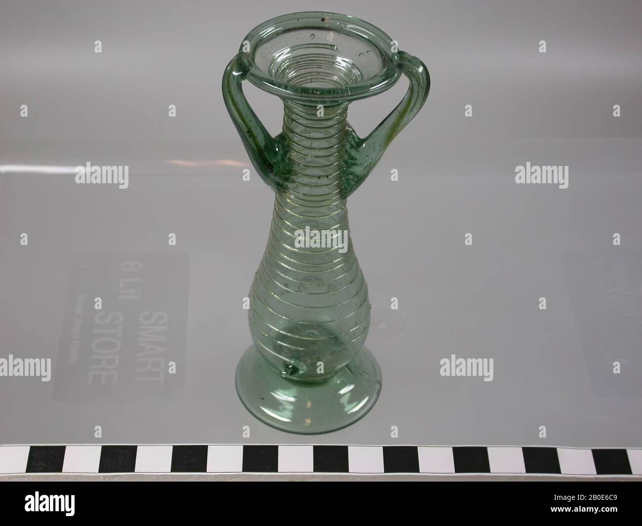 Bottiglia di vetro verde chiaro su un piede basso con un ventre convesso che termina in un collo stretto. Bocca larga con un orecchio verticale su entrambi i lati. Il corpo è avvolto a spirale con un filo di vetro, beffa, vetro, H 11,8 cm, Siria Foto Stock
