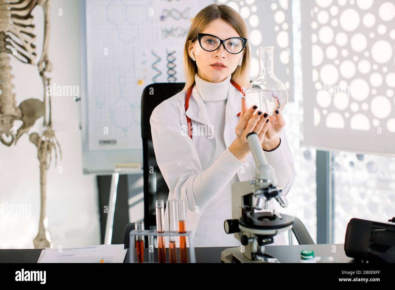 Una bella donna di ricerca medica o scientifica indossando cappotto bianco e bicchieri, contiene grande matraccio con liquido trasparente incolore, utilizzando Foto Stock