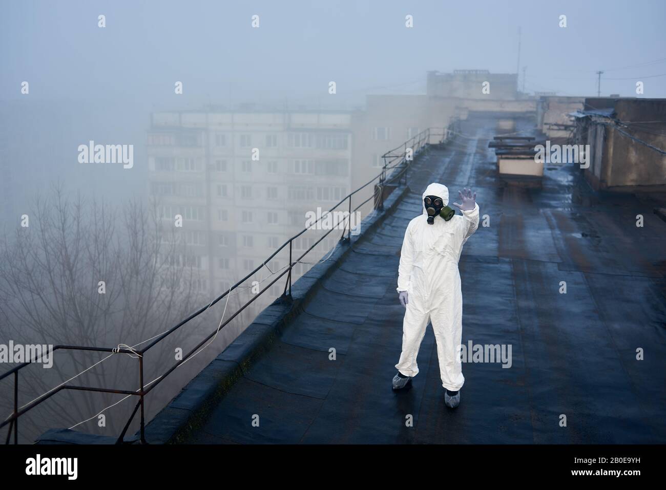 Ricercatore che si trova sul tetto di blocco di appartamenti, mostrando segno di stop, indossando tuta e maschera di protezione bianca, esplorando l'ambiente per la ricerca futura Foto Stock