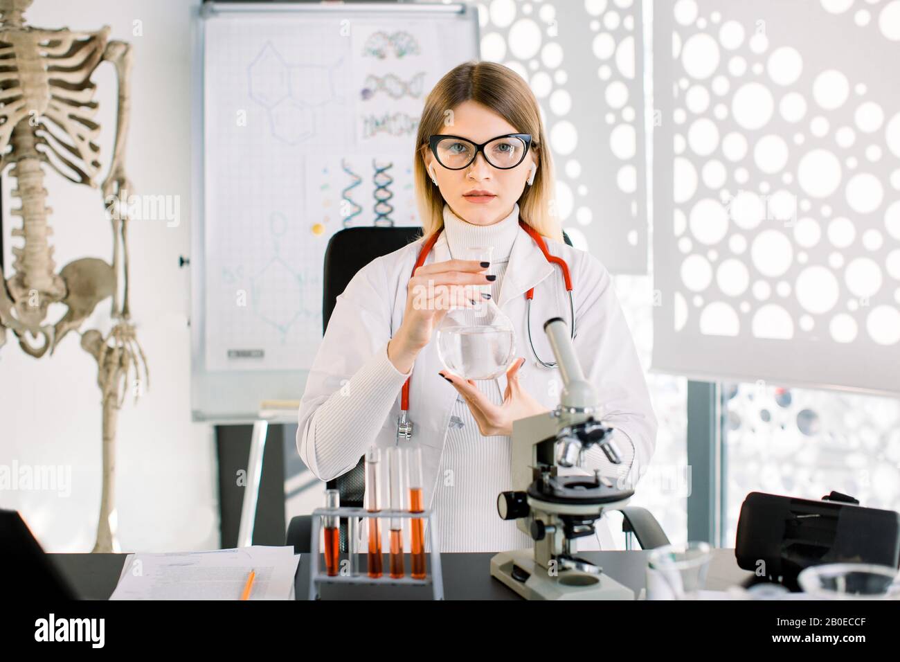 Smart grazioso scienziato caucasico giovane donna mostra matraccio di vetro con liquido all'interno, seduto al tavolo con microscopio e provette in Foto Stock