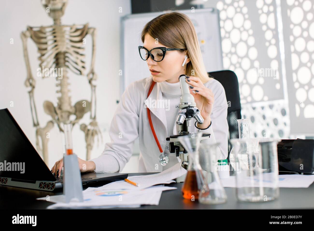 Bella donna caucasica scienziato in occhiali preparazione di un microscopio per test e analisi di laboratorio, prendendo appunti sul portatile. Laboratorio moderno Foto Stock