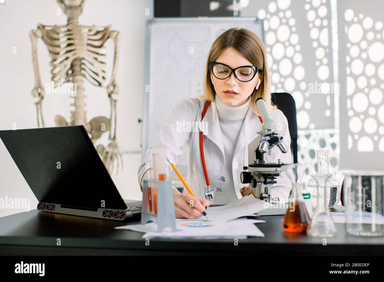 Giovane attraente scienziato caucasico donna in camice bianco e occhiali nel laboratorio chimico medico scientifico, seduto al tavolo con Foto Stock