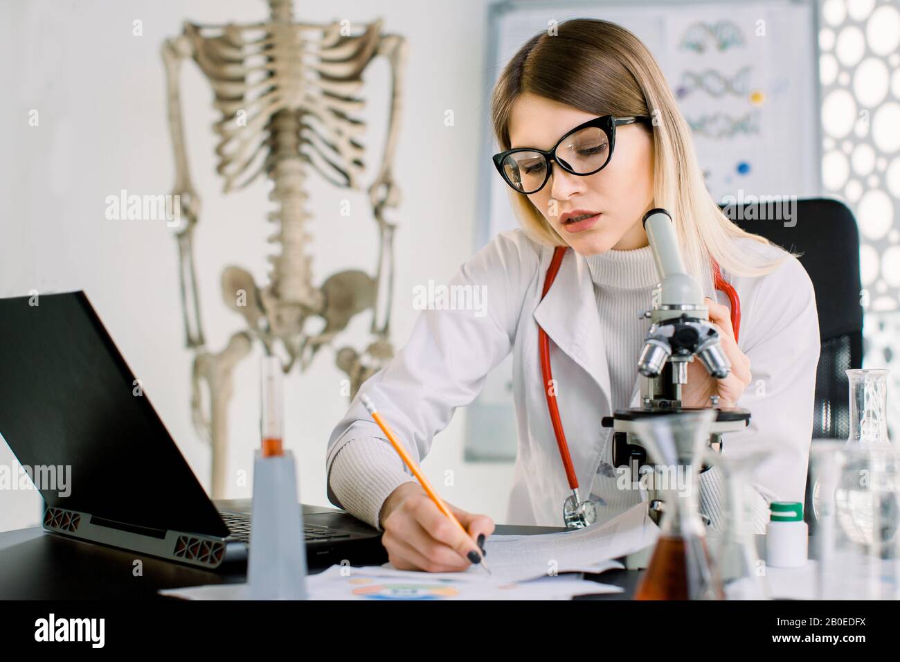 Giovane donna medico, biochimico, scienziato che guarda attraverso un microscopio in laboratorio, prendendo appunti sulle carte e sul laptop. Sala da laboratorio leggera Foto Stock