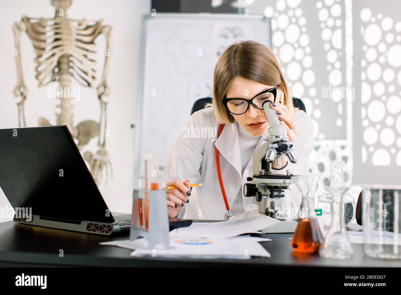 Bella donna caucasica medico scienziato in occhiali protettivi guardando al microscopio, seduto al tavolo con attrezzature da laboratorio e laptop Foto Stock