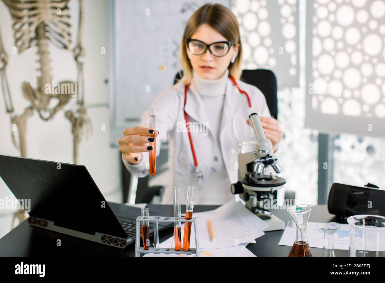 Giovane abbastanza caucasica donna scienziato guardando il campione scientifico, provetta con liquido rosso in mano, mentre seduto al tavolo con il portatile Foto Stock