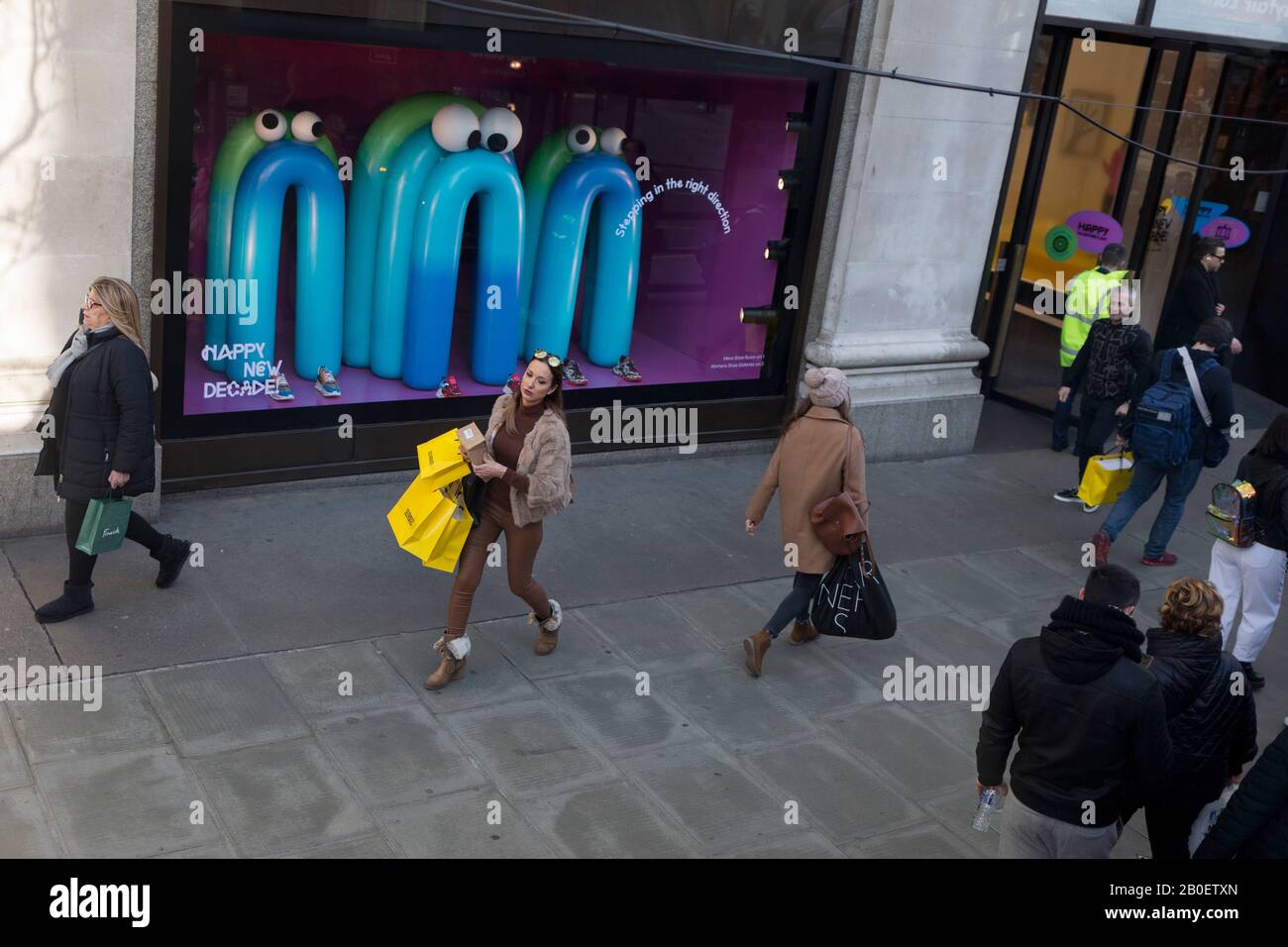 Portando con sé tre borse di shopping con marchio giallo, una signora emerge il grande magazzino Selfridges di Oxford Street a Londra, e passa da una vetrina a tema che include alcuni personaggi cartoon-esque che desiderano al pubblico un felice nuovo decennio, il 7th febbraio 2020, a Londra, Inghilterra. Foto Stock