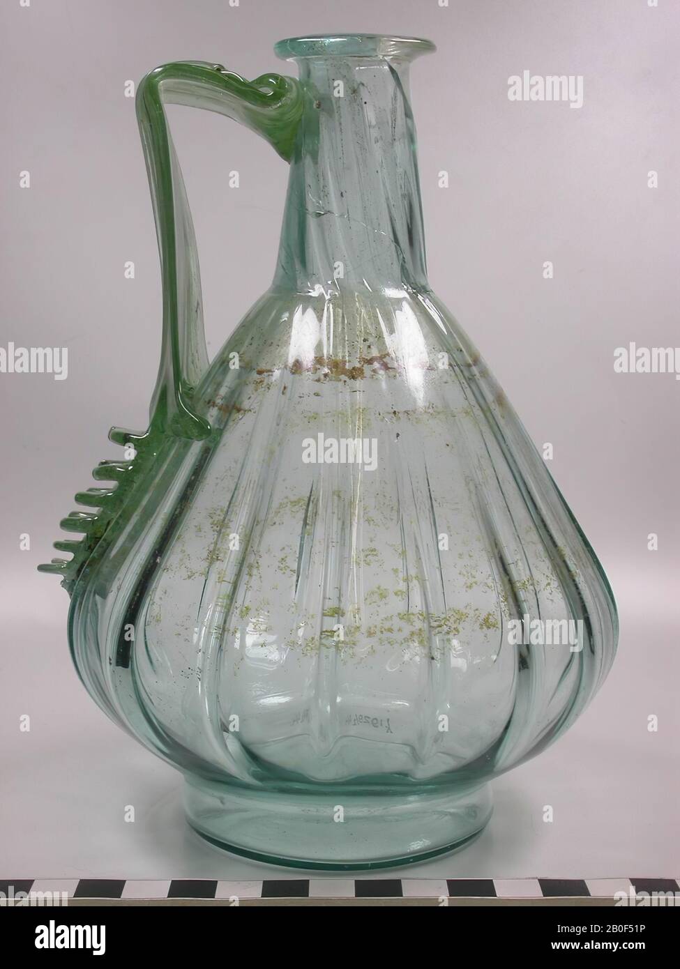 Caraffa in vetro da una tomba chiusa con numeri 17, 18 e 24. La brocca è parzialmente pre-soffiata in forma cilindrica e rifinita con tecnica di soffiatura libera. L'orecchio è acceso separatamente. Ad eccezione della frattura del labbro, questo oggetto è completamente intatto. Le creste leggermente attorcigliate sul collo sono causate da 90 gradi di rotazione rispetto all'addome durante la formazione. Il vetro è di colore blu-verdastro., beffa, brocca, vetro, altezza: 19,5 cm, roman 69-96 ad, Olanda, Brabante Nord, Cuijk, Cuijk Foto Stock