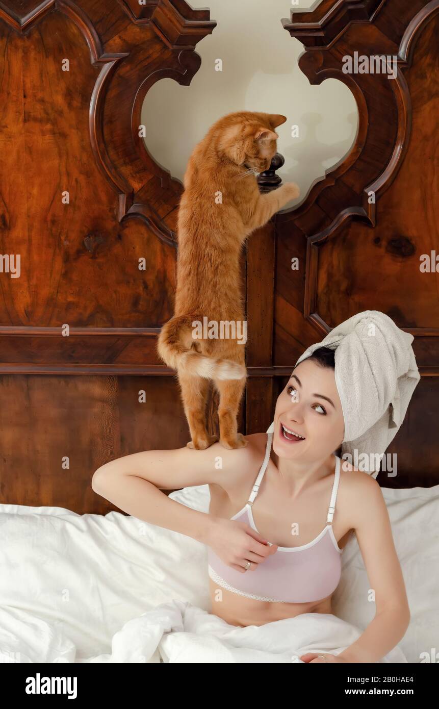 giovane donna con un gatto rosso sulla spalla, un asciugamano sulla testa si siede in un vecchio letto, sorridendo. Foto Stock