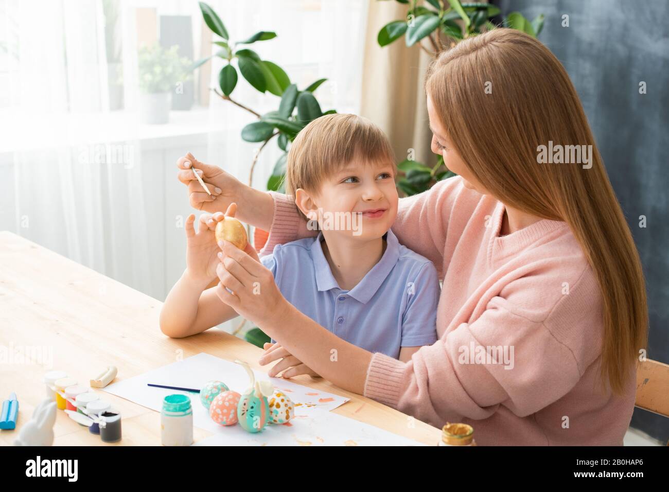 Figlio sorridente che guarda la madre mentre dipinge l'uovo di Pasqua in mano a casa Foto Stock