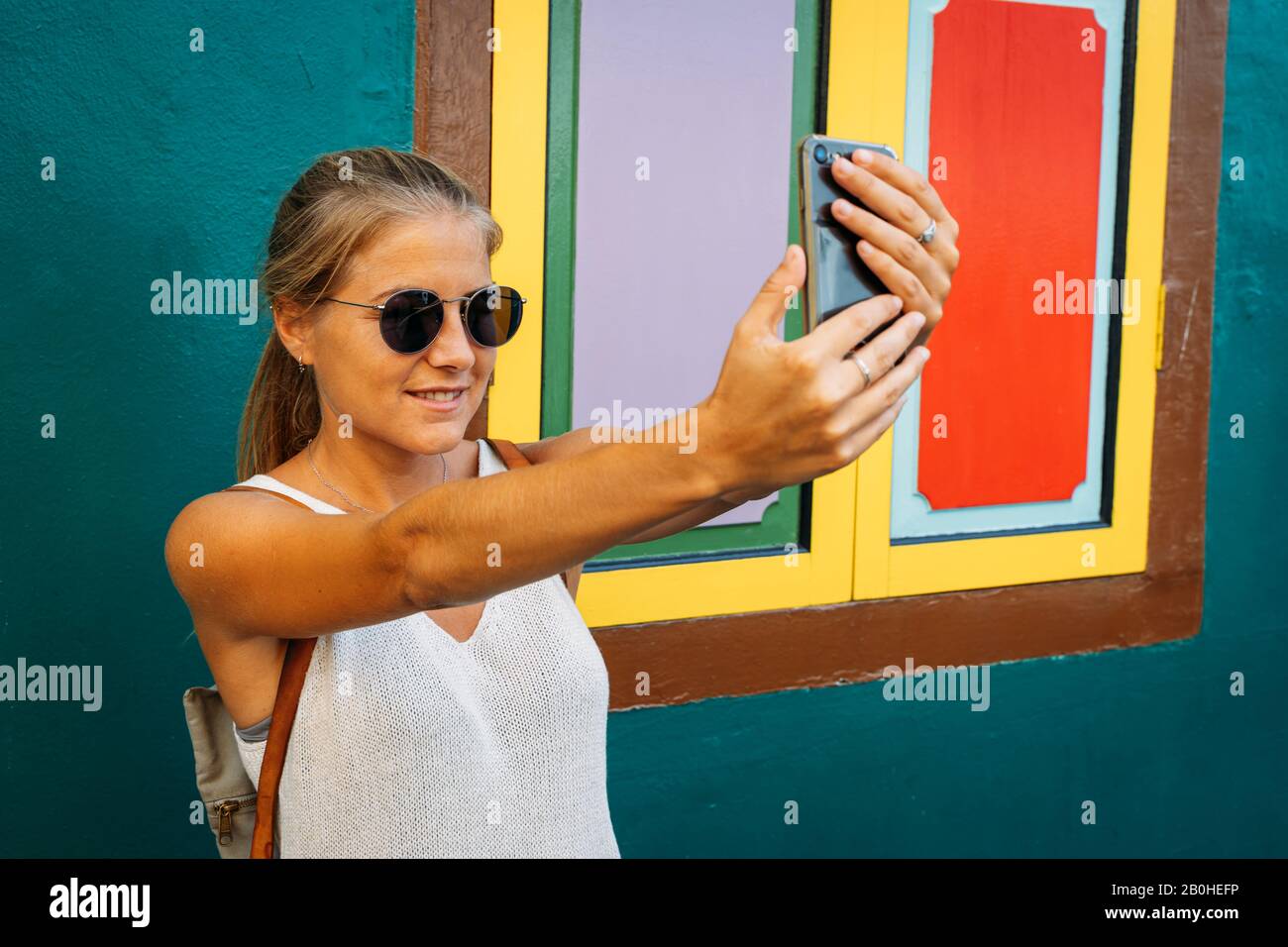 Ragazza bionda con occhiali da sole che fa un selfie con il telefono  cellulare di fronte a una finestra con i vetri colorati in una facciata  colorata Foto stock - Alamy