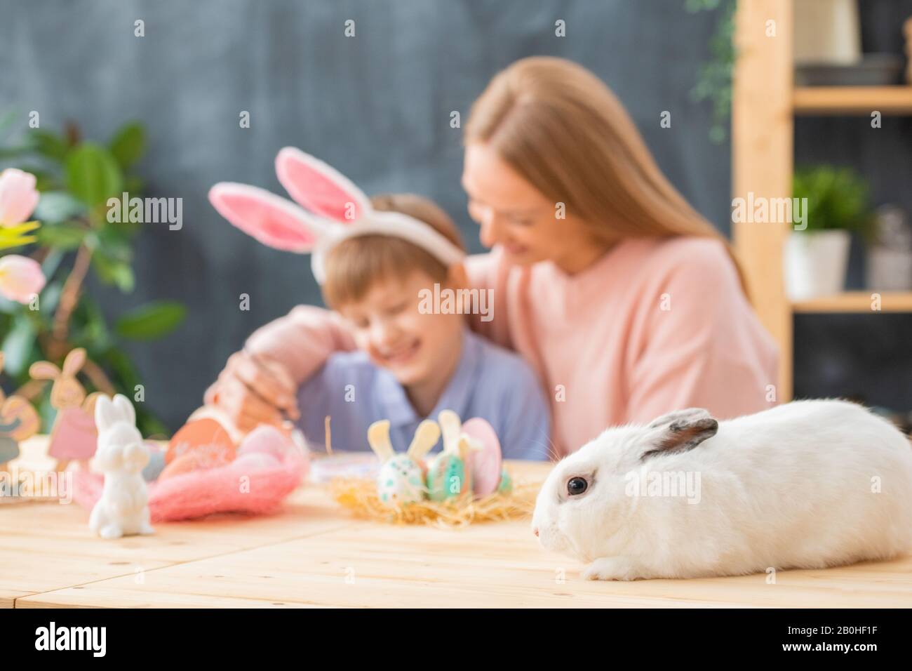 Primo piano di coniglio bianco soffice sdraiato su tavola di legno con decorazioni pasquali, madre che abbraccia figlio in background Foto Stock