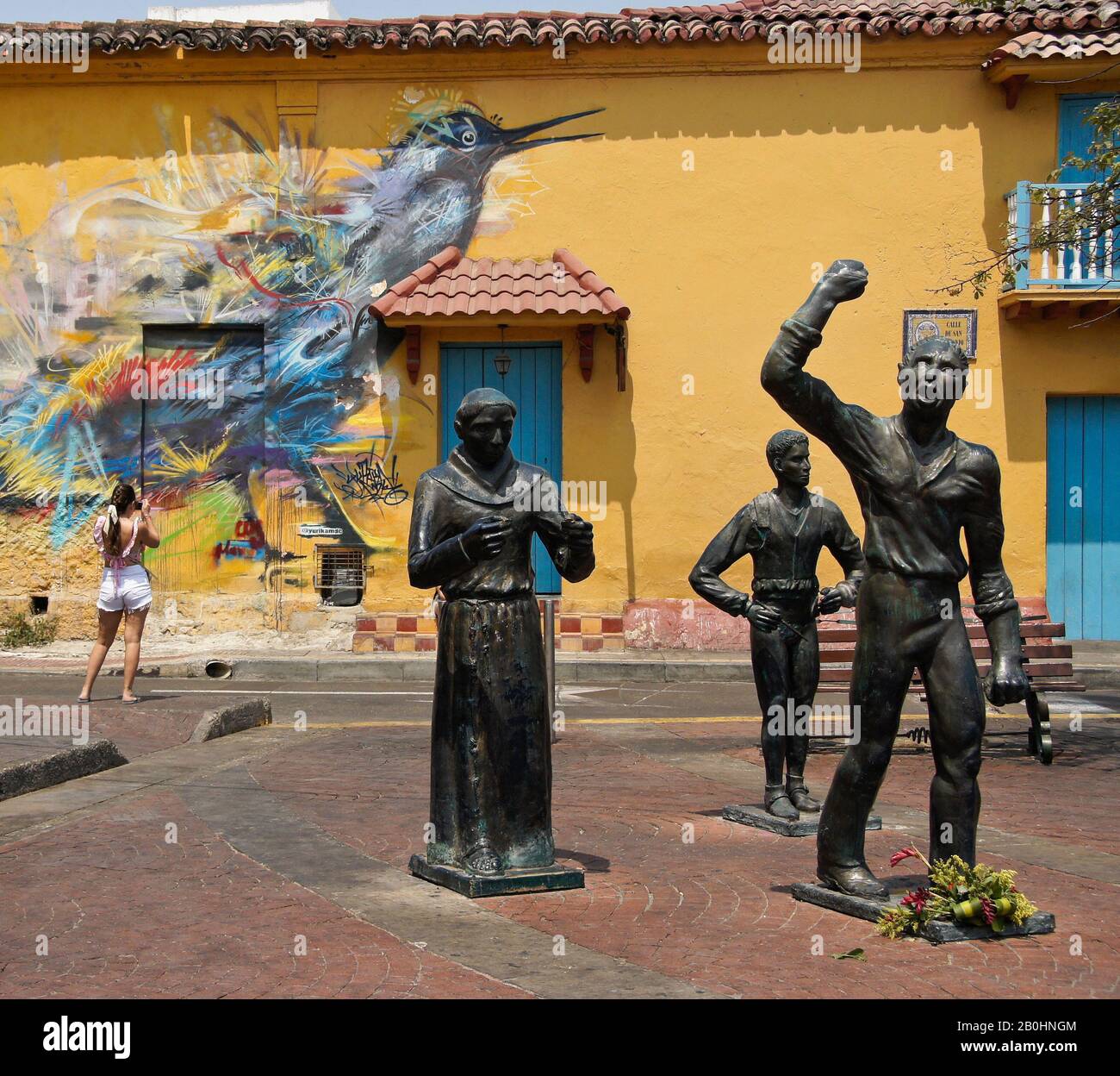 Sculture di eroi indipendenti in Plaza Trinidad di fronte all'arte di strada che decorano un edificio coloniale a Getsemani, Cartagena, Colombia Foto Stock