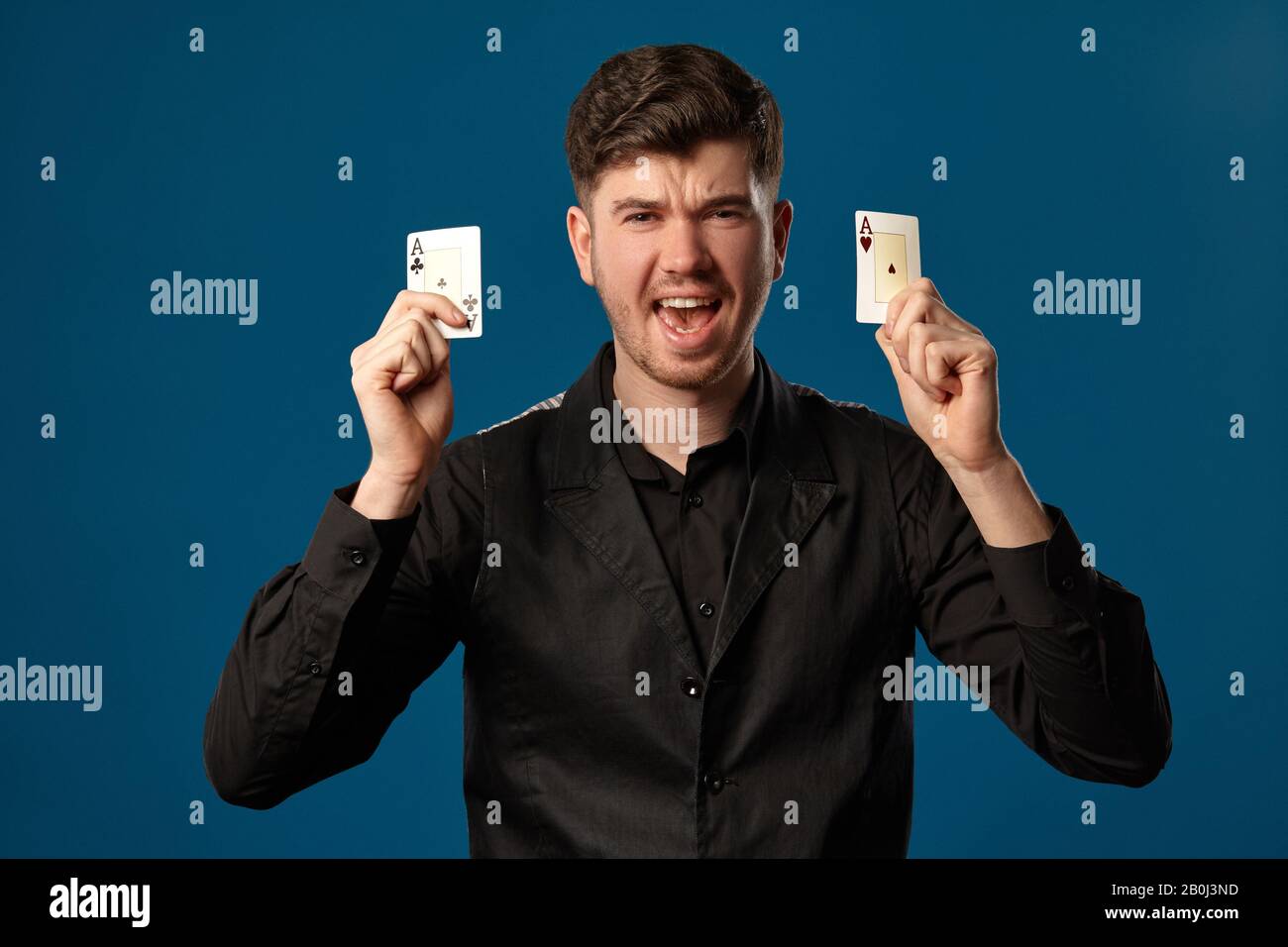 Newbie in poker, in gilet nero e camicia. Tenendo due carte da gioco mentre si posa contro sfondo blu studio. Gioco d'azzardo, casinò. Primo piano. Foto Stock