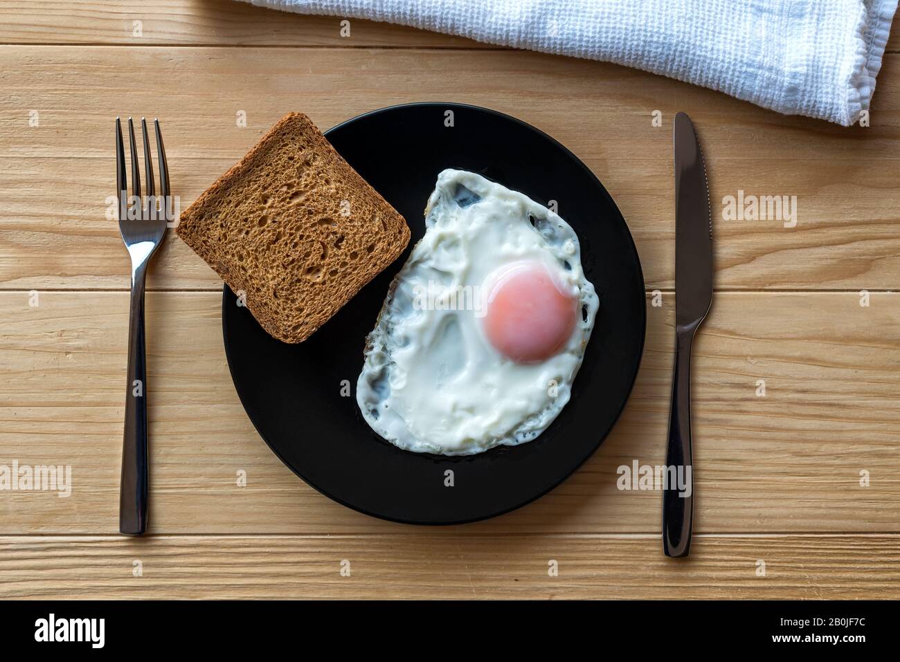 Uovo fritto chiudere in una piastra nera su un tavolo di legno con pane tostato e utensili. Cibo per la colazione. Lato soleggiato uovo fritto morbido Foto Stock