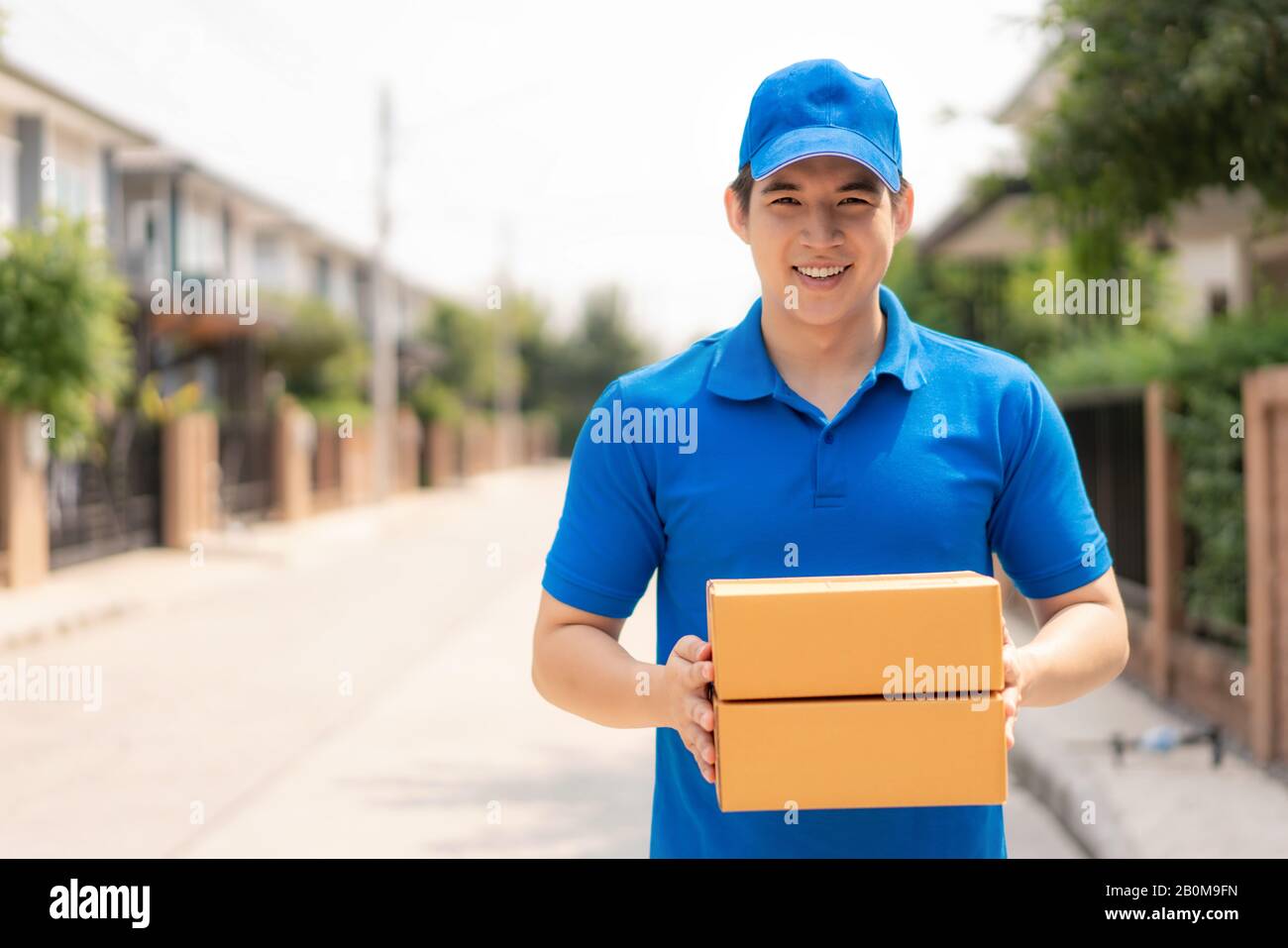 Asian consegna giovane uomo in blu uniforme sorriso e tenendo mucchio di scatole di cartone in casa villaggio di fronte con spazio copia. Pubblicità, Business, Tran Foto Stock
