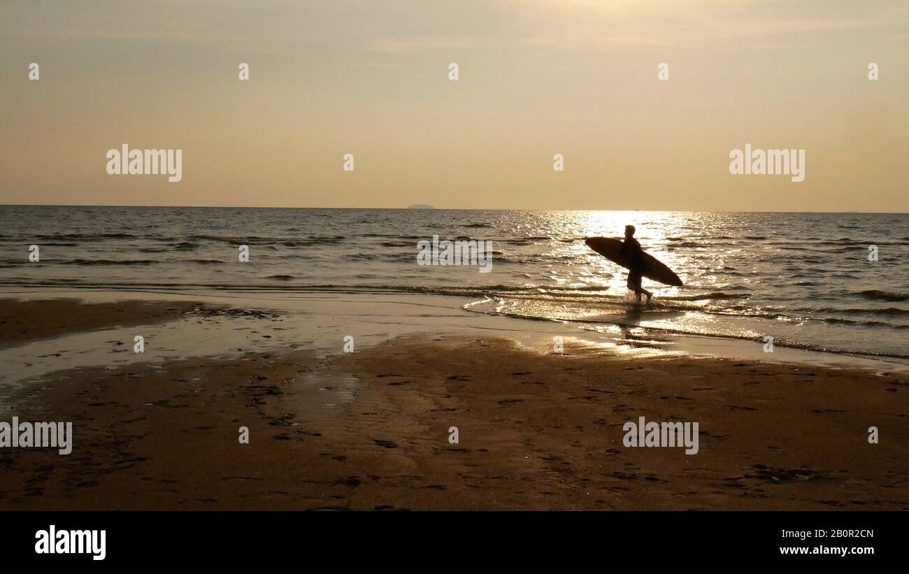 Silhouette di surf man con tavola da surf in esecuzione su superficie d'acqua. Surf alla spiaggia del tramonto. Sport acquatici all'aperto avventura lifestyle.Summer attività. Foto Stock