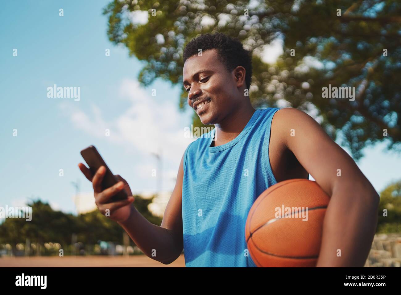 Ritratto di un giovane e sorridente giocatore di basket maschile che tiene la palla in mano messaggi di testo sul suo smartphone al parco Foto Stock