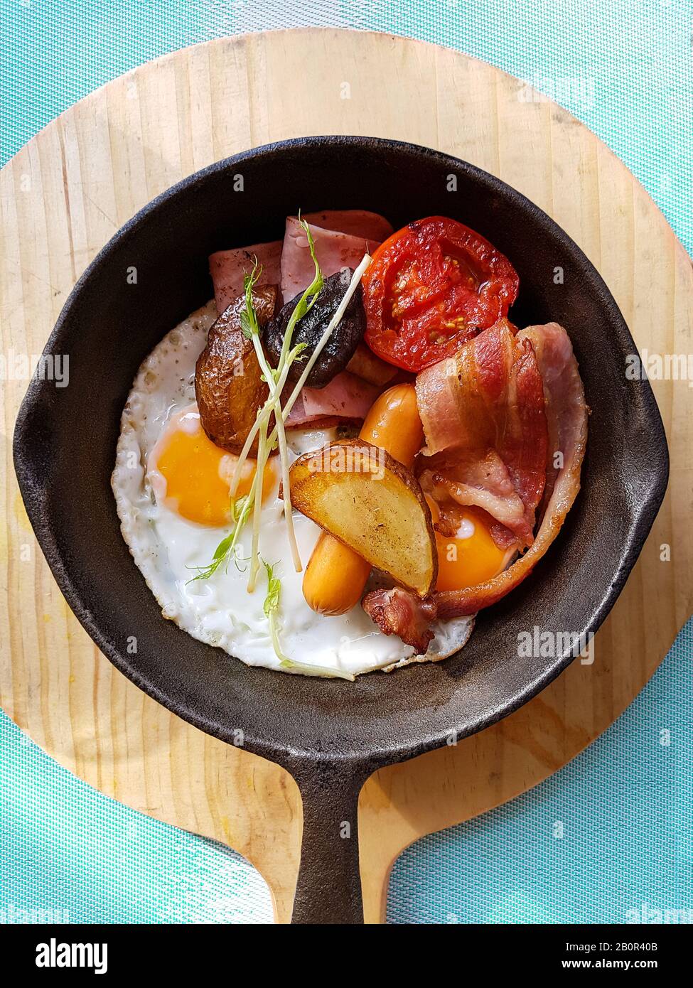 Prima colazione in padella con uova fritte, salsicce, pancetta, pomodoro, patate e verdure su fondo di legno sul tavolo in cucina. Foto Stock