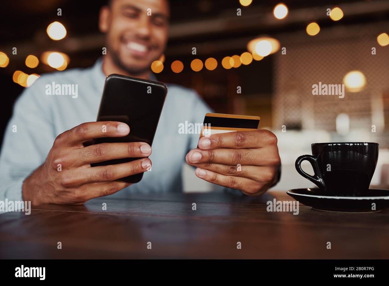 Closeup di allegro uomo africano mano che tiene il telefono cellulare digitando i dati della carta per effettuare il pagamento online seduto in caffetteria Foto Stock