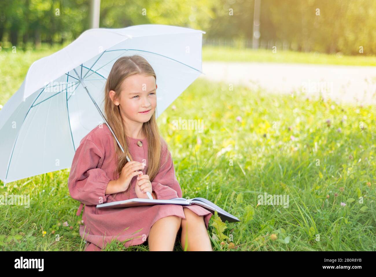 bambino che legge un libro. bambina sotto un ombrello si siede su erba verde in una luminosa giornata di sole Foto Stock