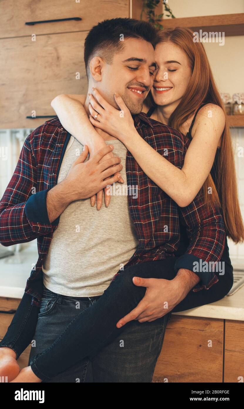 Bella ragazza caucasica con capelli rossi e frane abbracciando il suo amante, mentre in piedi sul tavolo in cucina Foto Stock
