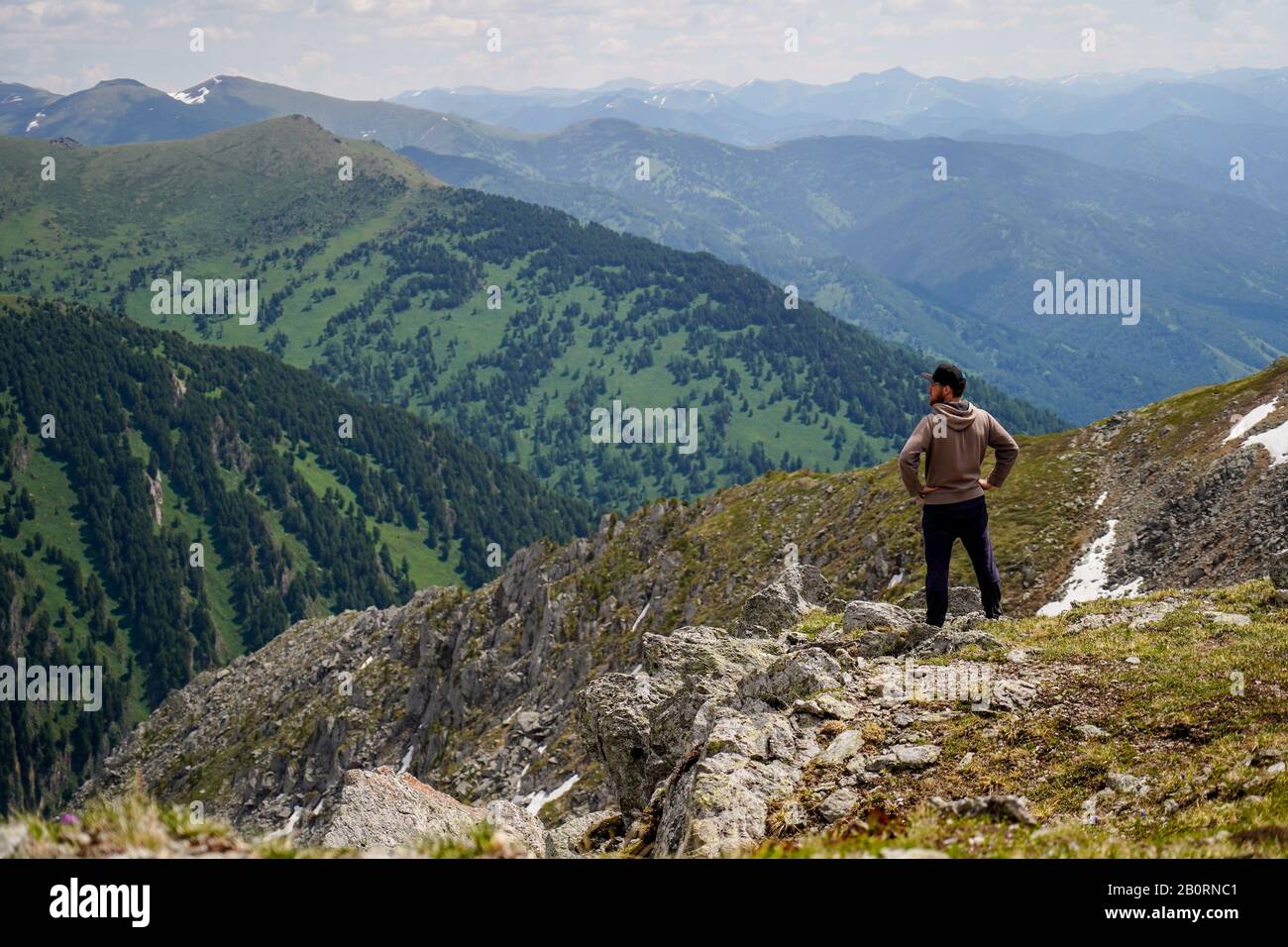 Un maschio con una felpa con cappuccio marrone, si voltò indietro sul pendio di montagna di fronte alla montagna sfondo Foto Stock