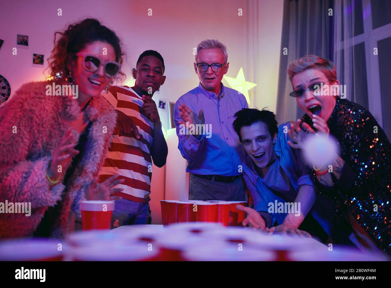 Uomo anziano che lancia la palla negli occhiali mentre gioca in partita a beer pong insieme ai suoi amici al party Foto Stock