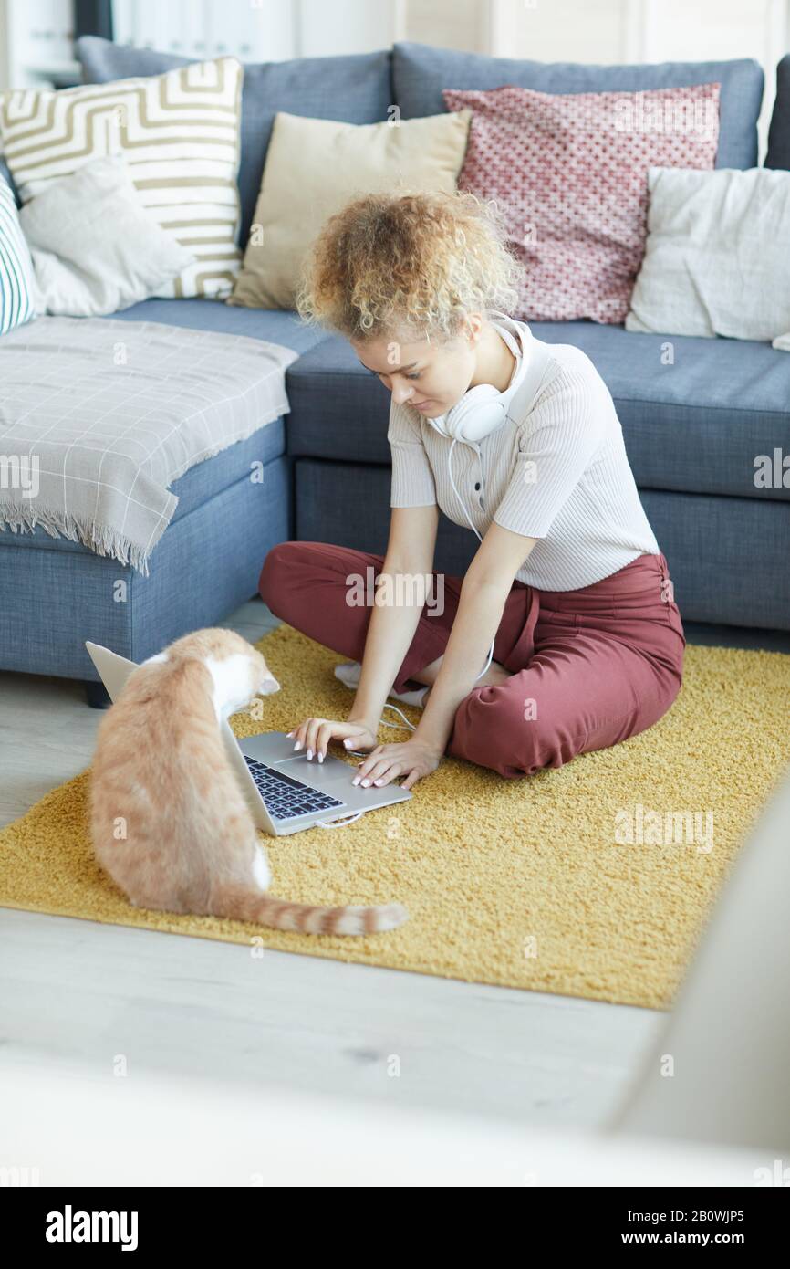 Giovane donna seduto sul pavimento che scrive sul suo computer portatile e gatto domestico che la disturba dal lavoro a casa Foto Stock