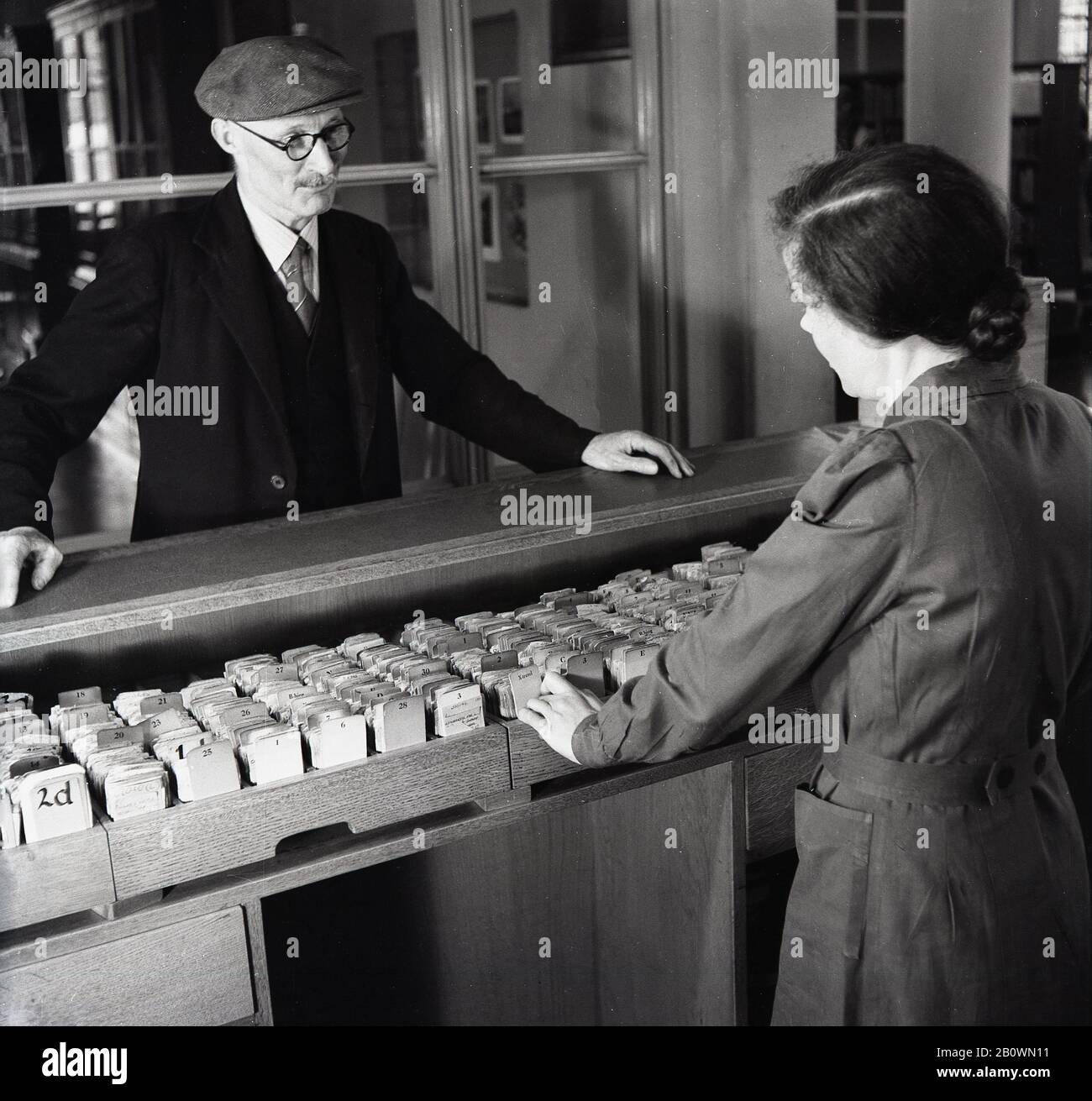 Anni '1950, anziano con camicia, cravatta e berretto piatto, che parla con una bibliotecaria femminile in piedi davanti a vassoi di legno di schede in questa foto storica di J Allan Cash. Foto Stock
