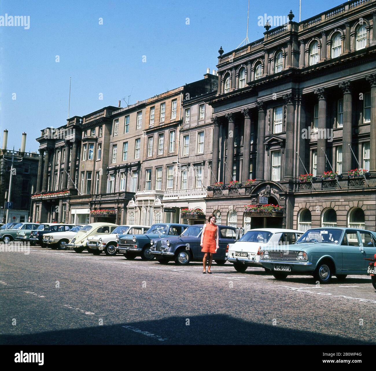 Alla fine degli anni '1960, storica, una signora con un vestito arancione in piedi accanto alle auto dell'epoca, parcheggiata fuori dal George Hotel, George Street, Edimburgo, Scozia, Regno Unito con auto dell'epoca parcheggiate all'esterno. Foto Stock